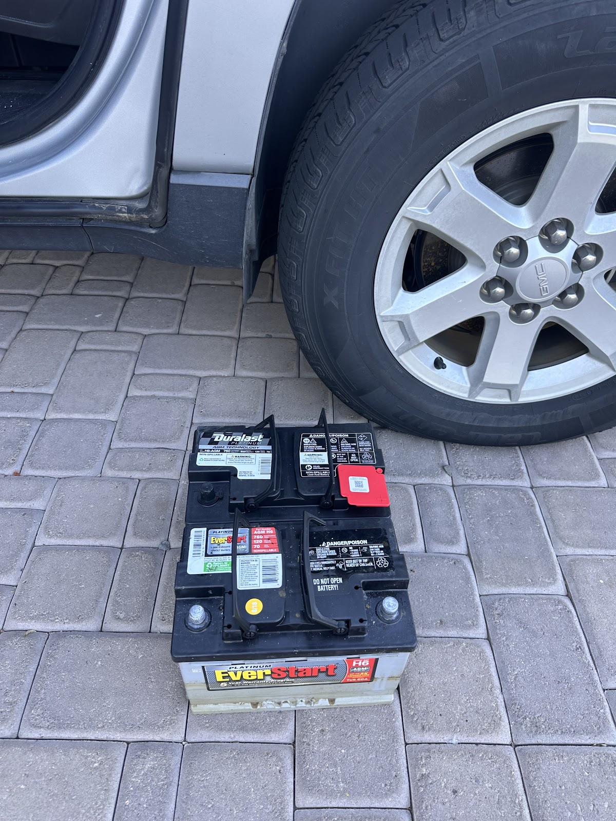 The old battery stacked on the new replacement battery on paver stones beside the wheel