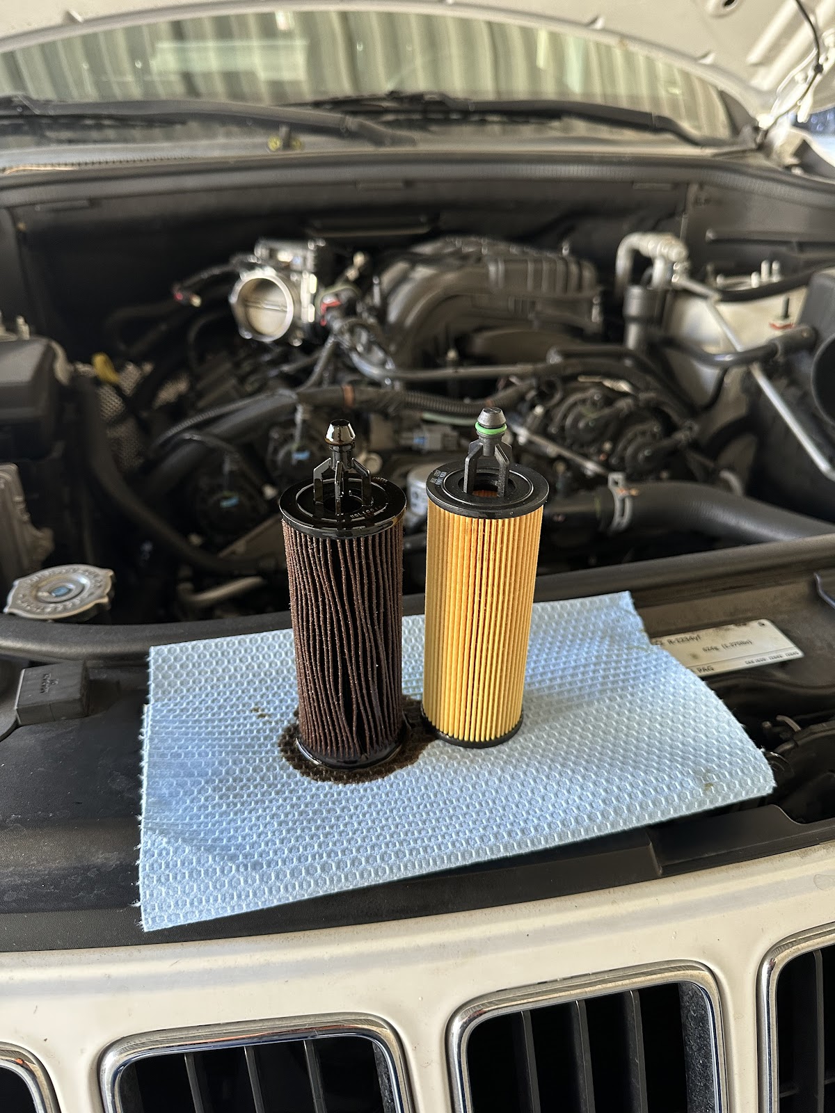 Dirty old oil filter standing next to the clean new one on a shop towel over the engine bay